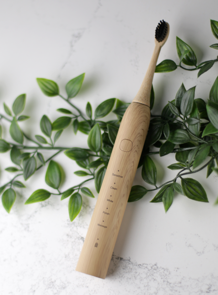 A eco sustainable bamboo electric toothbrush made of recyclable material is sitting on a marble countertop on top of green leaves 