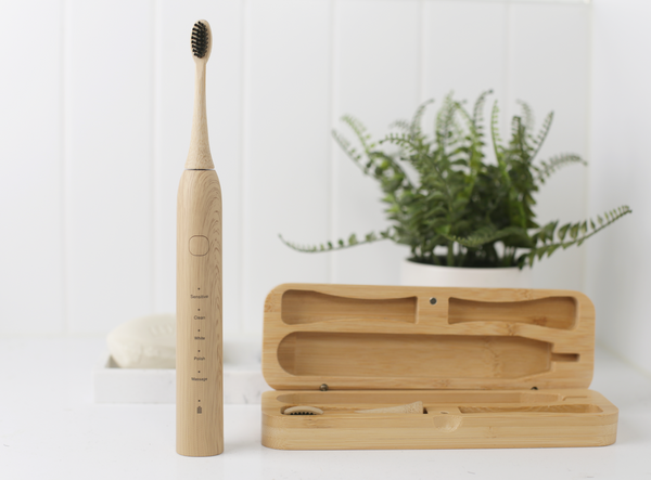 A eco sustainable bamboo wooden electric toothbrush is standing upright next to its bamboo toothbrush case. Both items are displayed in a white bathroom  in front of a white tiled wall